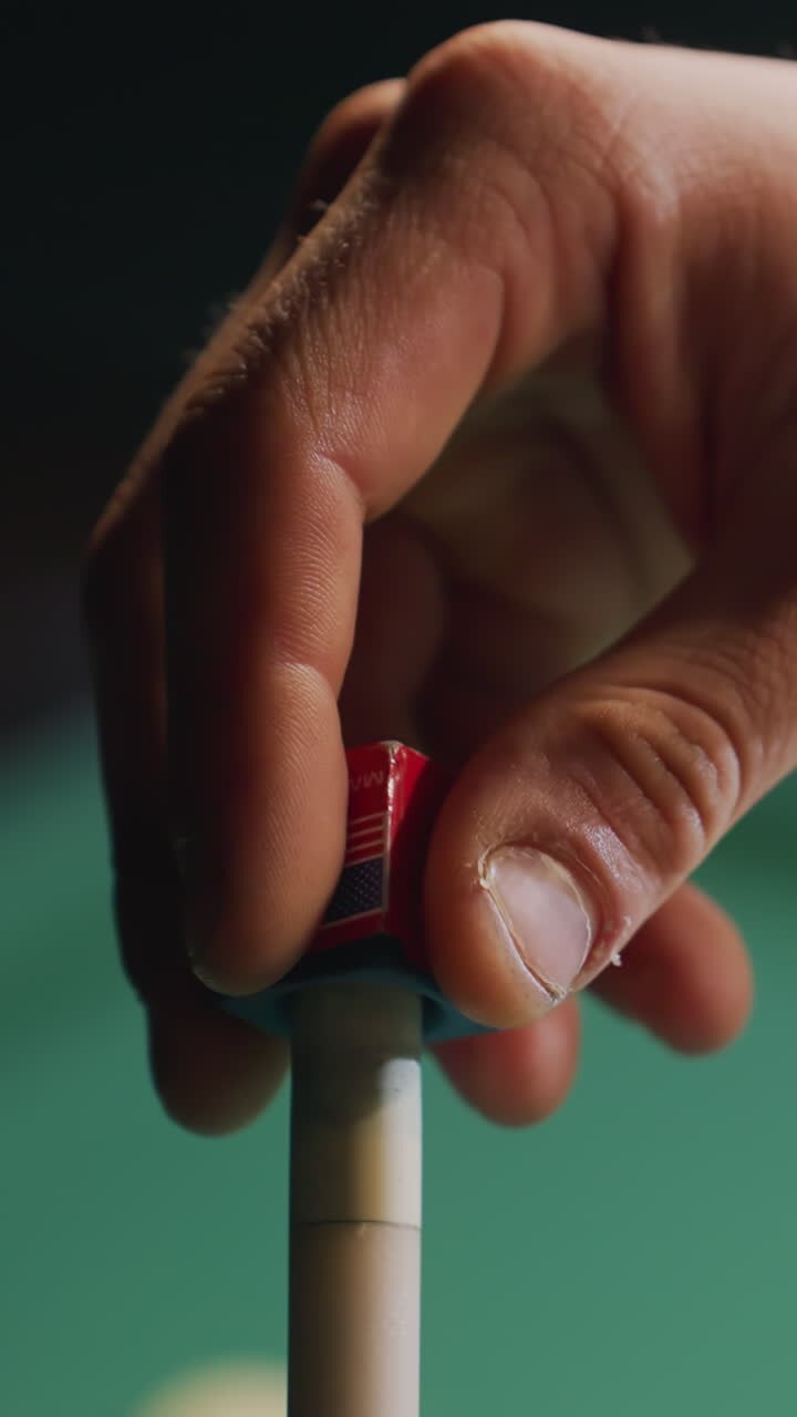 Close-up of player's hand applying chalk to cue stick before executing precise shot on green pool table. Focus on preparation and technique with blurred billiard balls in background