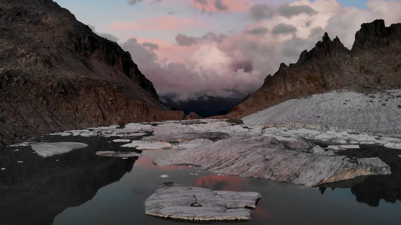 sobrevuelo aéreo giratorio sobre un lago glaciar lleno de icebergs derretidos en partes remotas de los alpes suizos durante la puesta de sol con una panorámica desde el reflejo del horizonte hasta la parte superior de los trozos de hielo