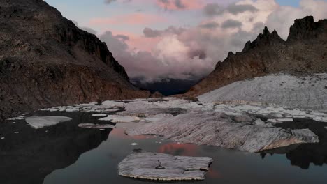 Drehende-Luftüberführung-über-Einem-Gletschersee-Voller-Geschmolzener-Eisberge-In-Abgelegenen-Teilen-Der-Schweizer-Alpen-Während-Des-Sonnenuntergangs-Mit-Einem-Schwenk-Von-Der-Horizontreflexion-Nach-Unten-über-Die-Eisbrocken