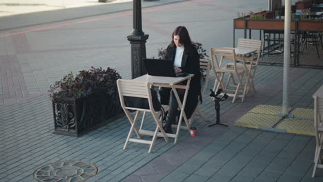 young woman working on laptop outdoors at a restaurant surrounded by wooden chairs, plants, and a tripod, she is seated at a table with a coffee cup, with a paved walkway and blurred background