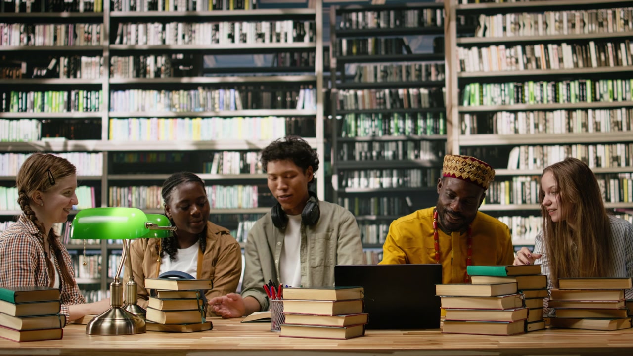 Gruppo di studio in biblioteca che discute idee attorno a un laptop e libri impilati