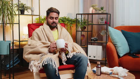 sick indian man wrapped in plaid sit alone shivering trembling from cold on chair drinking hot tea