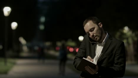 hombre de negocios usando teléfono inteligente hacer una llamada mientras está de pie en el parque en una plaza de la ciudad, y hacer notas
