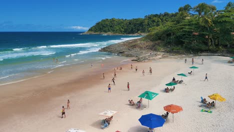 aerial over praia da tiririca beach with colorful umbrellas, people playing altinha, blue ocean, green jungle - itacare, bahia