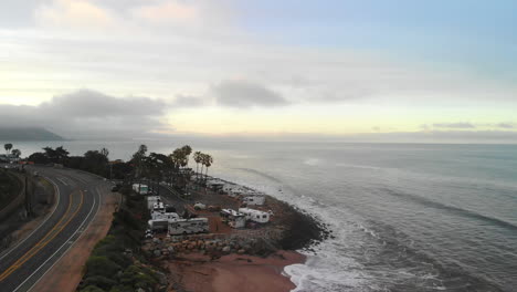 Drone-Aéreo-Alejándose-De-Un-Campamento-De-Rv-Y-Carretera-En-Las-Playas-De-Arena-De-Ventura,-California-Con-Olas-Oceánicas-Rompiendo-En-La-Orilla-Al-Amanecer