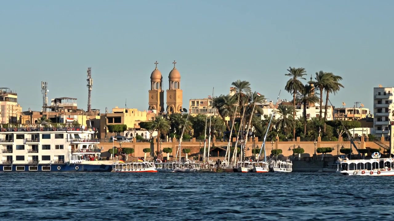 Un'ampia vista dal fiume Nilo che cattura l'argine di Luxor, con navi da crociera ormeggiate, barche tradizionali, imponenti palme e le prominenti guglie gemelle di una chiesa sotto un cielo azzurro e limpido