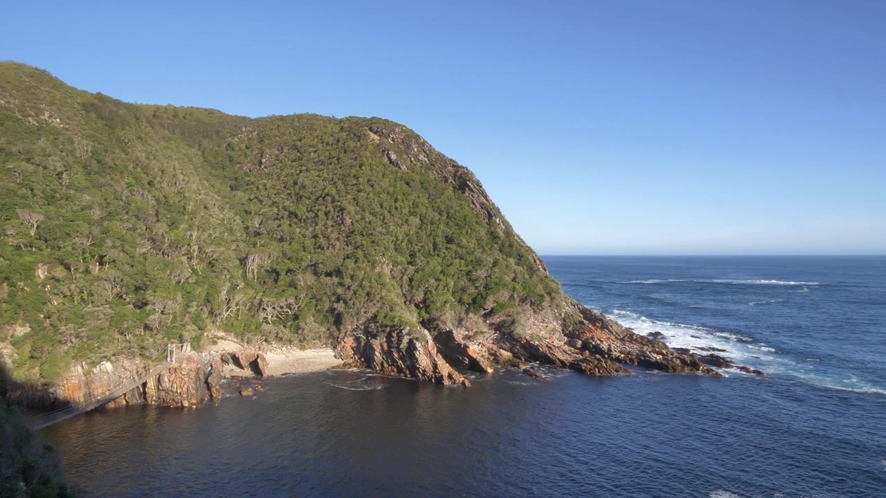 Vista panoramica di aspre scogliere costiere ricoperte da una fitta vegetazione verde, con onde oceaniche di un blu profondo che si infrangono contro la costa rocciosa e un ponte sospeso che collega elegantemente due punti a Tsitsikamma.