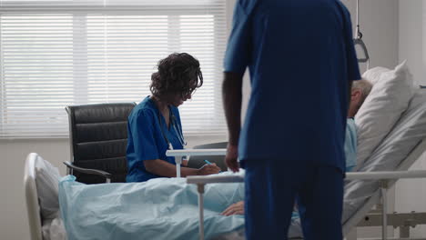 african young doctor team consulting covid-19 infected patient in bed at hospital. sick man lying in hospital bed and talking to surgeon in scrubs and protective mask. doctor examining elderly patient