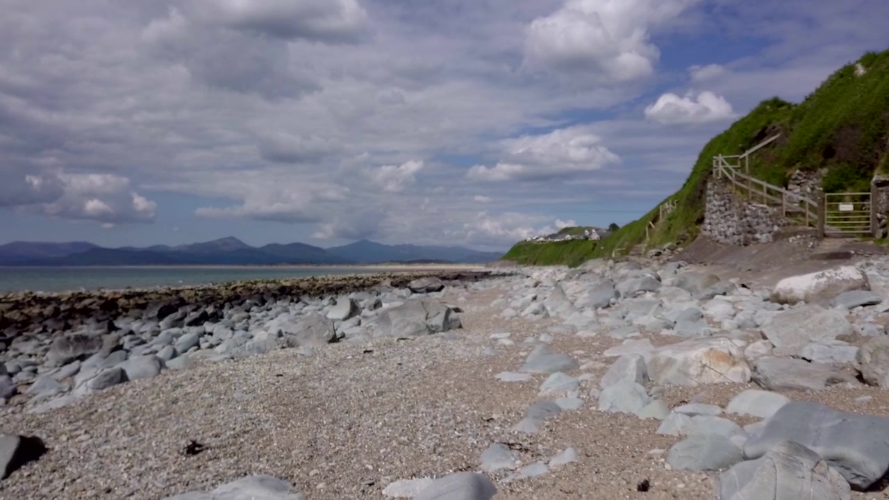 Un lent mouvement de drone fluide de droite à gauche le long d'une plage rocheuse vers des rochers plus sombres, avec une montagne lointaine à l'horizon, au Pays de Galles, au Royaume-Uni - version de 20 secondes.