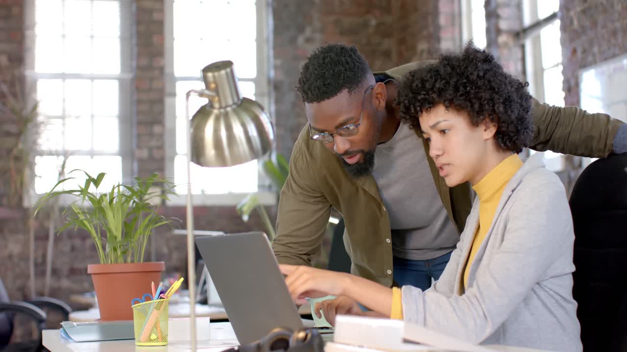 Focused diverse colleagues discussing work at table and using laptop in office in slow motion