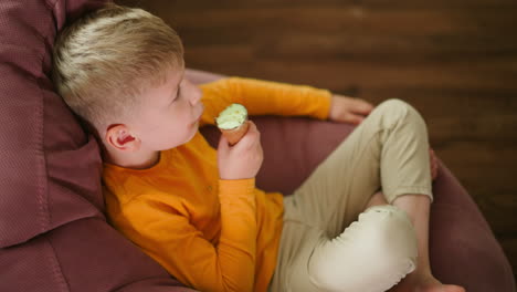 young boy in orange top seated barefoot on soft pink beanbag eating ice cream cone while a drop of ice cream falls on shirt, glancing at the mess with innocent expression