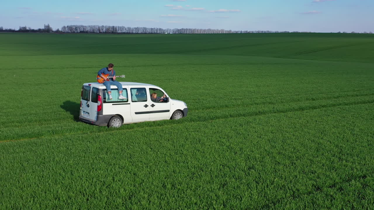 Young man playing the guitar on a moving car. Friends in white car travel on green field while guitarist sitting on a roof of the auto. Extreme for young people.