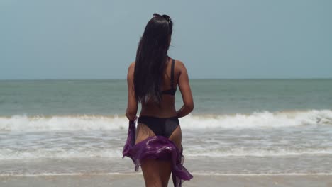 A-young-girl-in-a-black-bikini-walks-out-to-the-ocean-waves-crashing-at-the-shoreline-holding-a-shaul