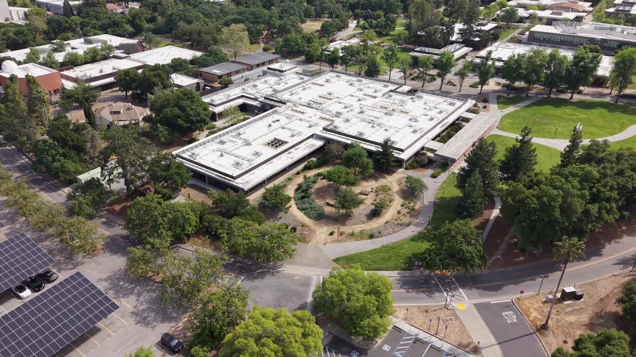 Imágenes aéreas de dron del campus de West Valley College en Saratoga, California. Se ve un gran edificio académico, rodeado de exuberantes árboles verdes, senderos y un estacionamiento con paneles solares.