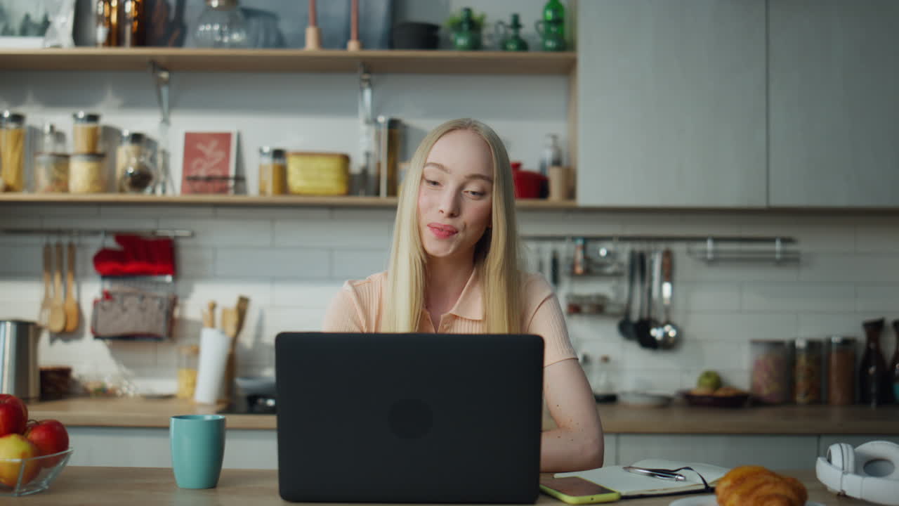 Girl chatting web camera sitting at kitchen table. Woman waving hand greeting.