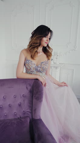 Fantastic young woman in a luxury dress is posing to camera. Beautiful model is standing behind the royal armchair in a long wedding dress on the background of studio. Vertical video