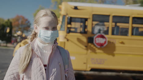 child in a protective mask stands in front of a school bus. a stop sign is visible at the rear.