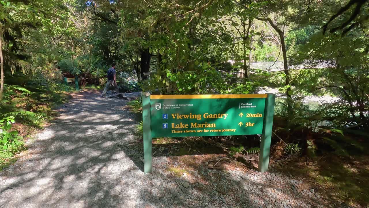 A man walks past a sign identifying the start of the track leading to Lake Marian in New Zealand. This beautiful hiking trail can be found in the Fiordland National Park in the South Island