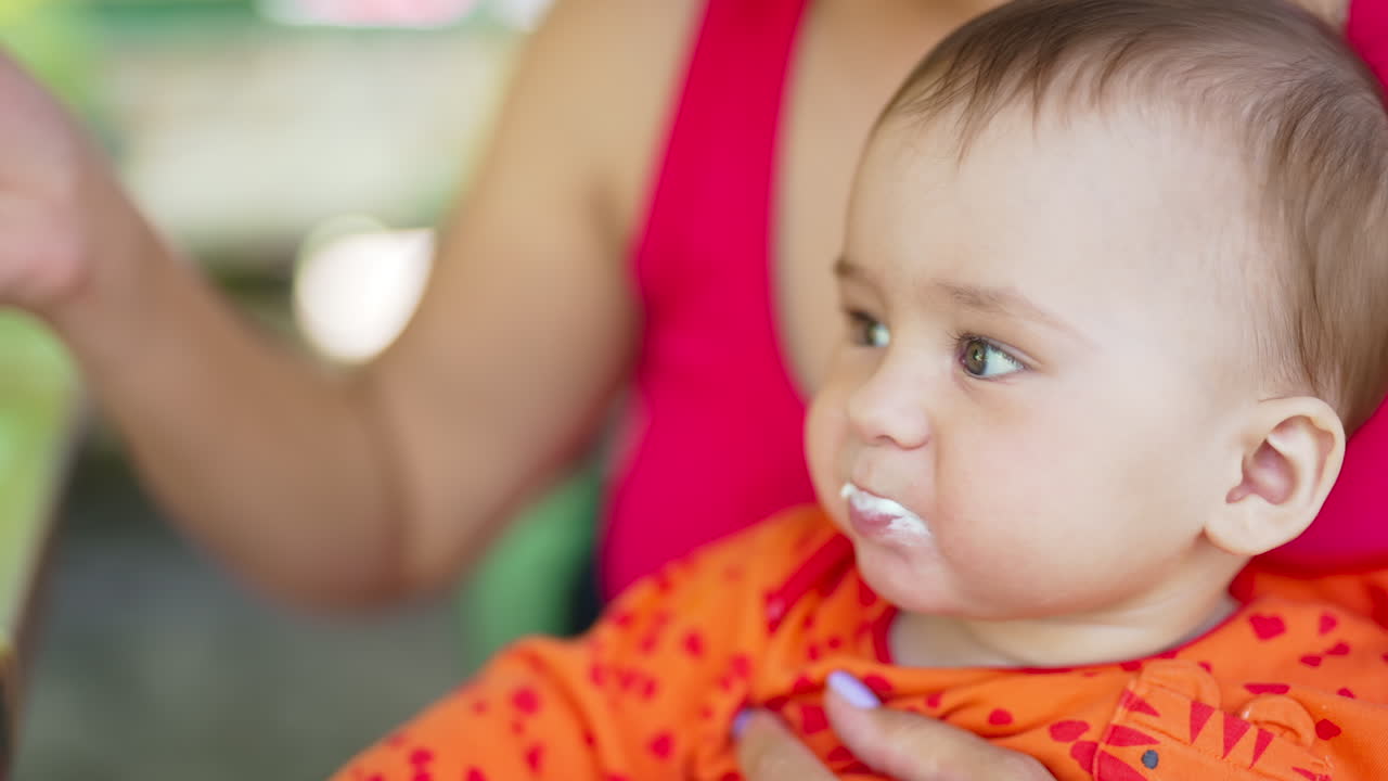 Cute infant being fed from a spoon. Mommy gives her child sitting on her laps tasty yoghurt. Close up.