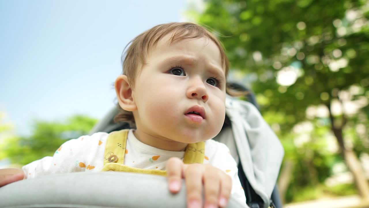 진지한 표정으로 유모차에 앉아 있는 동안 놀이터에서 노는 다른 아이들을 쳐다보는 호기심 많은 어린 소녀, 슬로우 모션, 낮은 각도로 얼굴 클로즈업