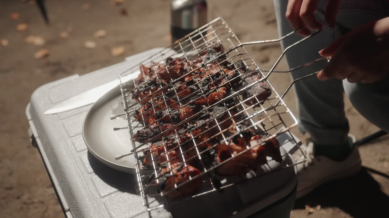 Tusuk sate dibalik di atas kotak pendingin di samping kursi, tangan menyesuaikan rak kawat dengan penjepit, koki aktif menyiapkan kebab di atas kotak pendingin busa, daging mendesis dan asap, alas kaki kasual luar ruangan dan celana jins terlihat, dinamis