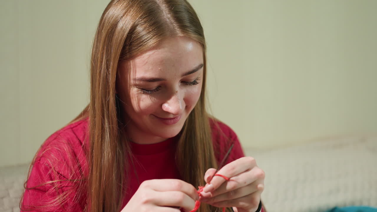털실로 공예를 하는 행복한 여성, 빨간 실로 작은 스티치를 즐겁게 만드는 젊은 여성, 밝은 실내 조명 아래에서 꼼꼼하게 뜨개질을 하면서 미소짓는 여성의 기분 좋은 클로즈업