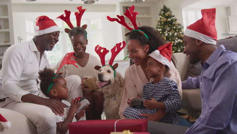 Multi-Generation-Family-Celebrate-Christmas-At-Home-Wearing-Santa-Hats-And-Antlers-Opening-Presents