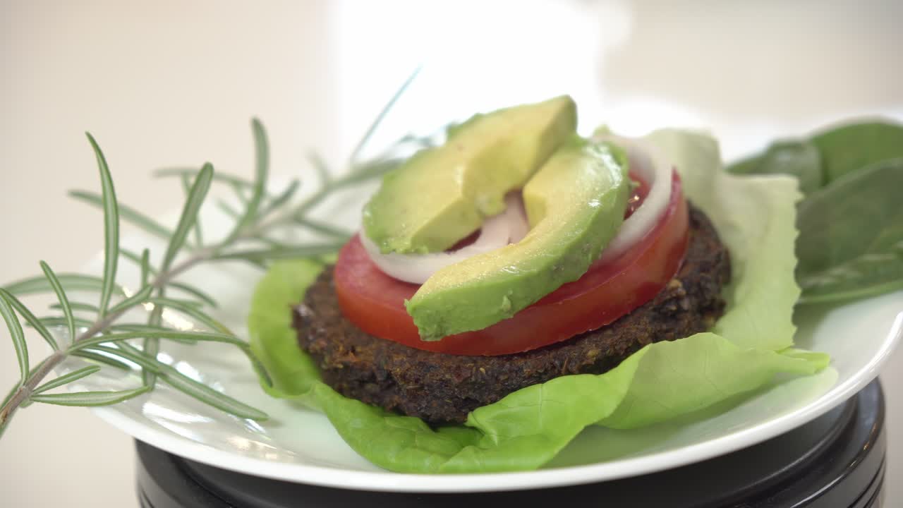 un bel shot di hamburger vegano guarnito con timo, fette di avocado, foglie di spinaci e lattuga al burro ruotando su un piatto.