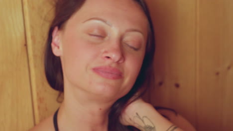 close up of cheerful woman with green eyes smiling warmly while resting hand on head, gazing directly into camera in serene sauna setting, soft lighting highlighting natural expression