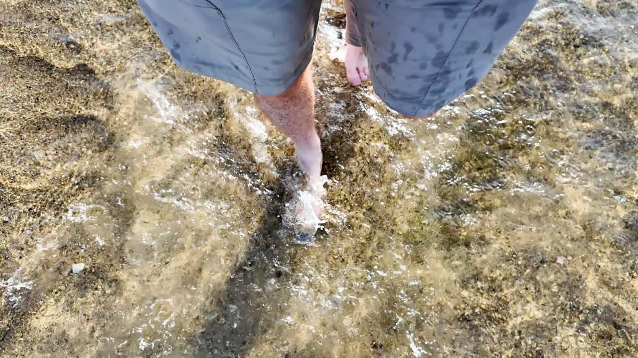 man walking bear foot in the sand while water is splashing over his feet in the ocean. The angle is top down on his feet while he's wearing tan shorts