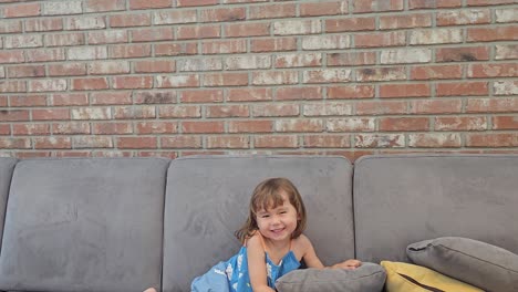 Little-girl-playfully-jumping-on-couch,-smiling-and-laughing,-looks-at-camera