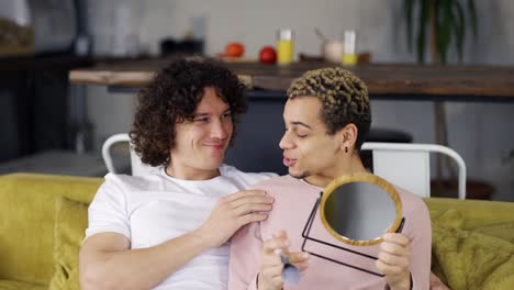 two gay friends sitting on the couch, a man doing make up with brush and mirror