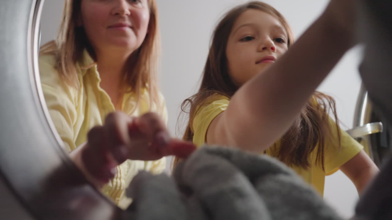 Inside washer pov shows laundry worker with little girl reaching in, removing fabrics toward lens, soft focus cloth, teamwork during wash cycle, family help, clean textiles, yellow shirts visible