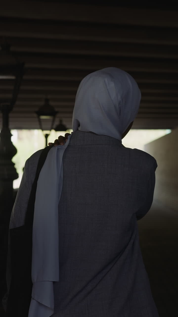 Muslim woman in grey coat walks along empty embankment under overpass bridge backside view slow motion. Muslimah in old city. Arabic traditions