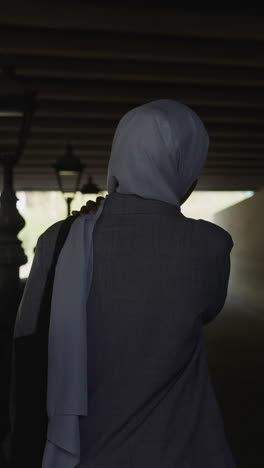 muslim woman in grey coat walks along empty embankment under overpass bridge backside view slow motion. muslimah in old city. arabic traditions