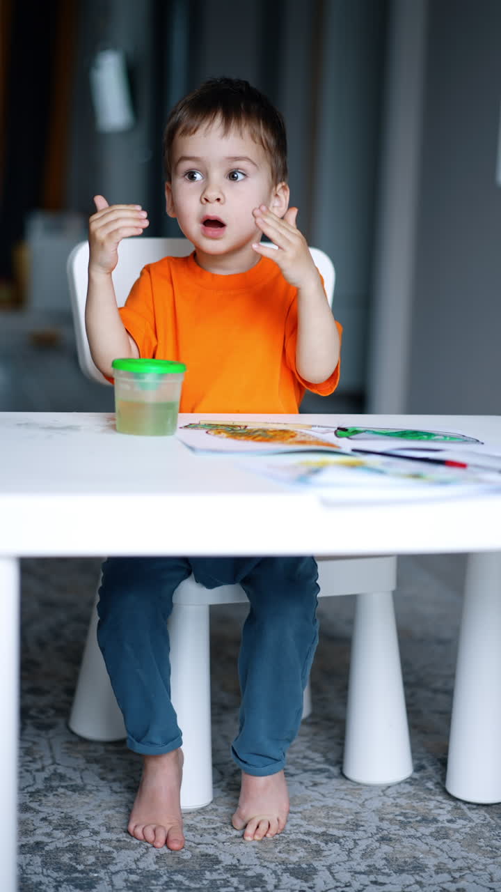 Little barefoot toddler sits at the desk clapping hands. Baby boy is getting ready to paint. Black cat walks around the kid. Vertical video.