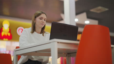 trabajador remoto sentado en un restaurante moderno trabajando en una computadora portátil, mirando hacia otro lado pensativo mientras toma un descanso, con un interior colorido y una iluminación vibrante en el fondo