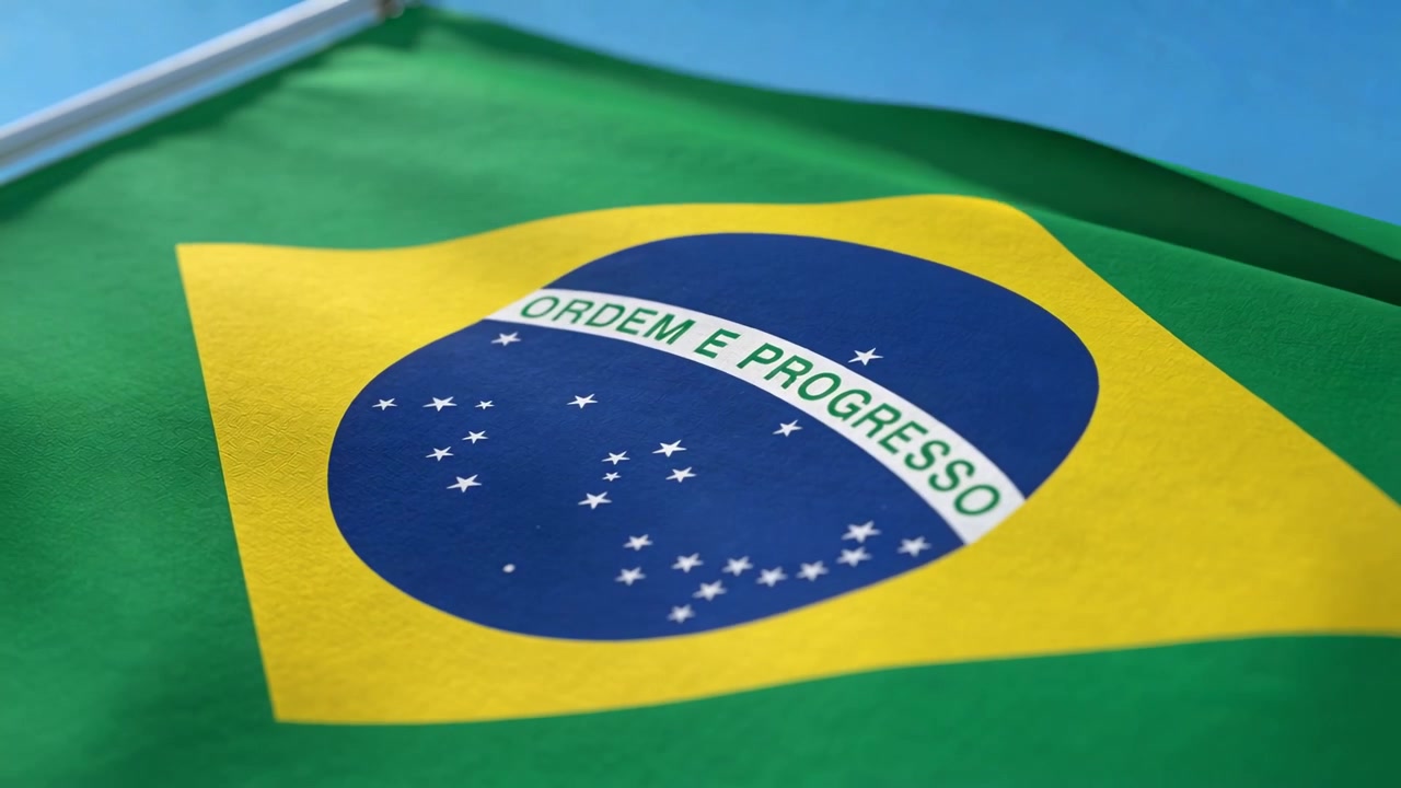 Vajende brasiliansk flag på stang afslører ORDEM E PROGRESSO-emblemet mod blå himmel i let brise.