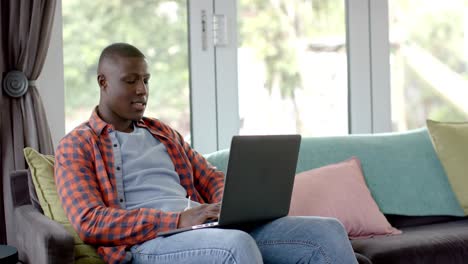 feliz hombre afroamericano sentado en el sofá usando computadora portátil en casa, cámara lenta