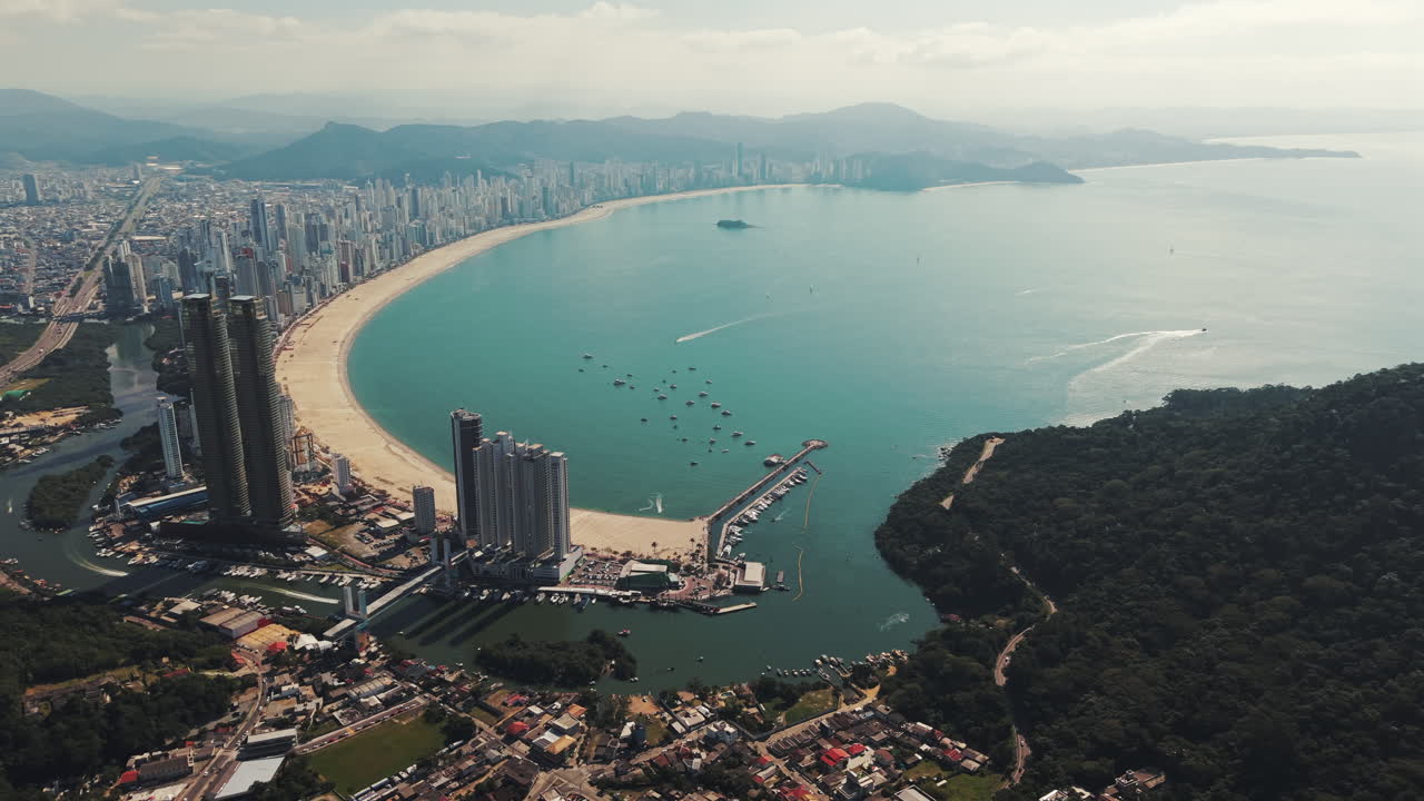 Smooth aerial drone orbit showcases the sweeping arc of Balneário Camboriú’s beach, turquoise bay, and impressive high-rise skyline set against lush green hills in southern Brazil