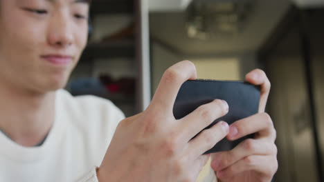 헤드폰을 착용 한 아시아 소년 이 집 의 소파 에 앉아 스마트폰 에서 게임 을 하고 있다