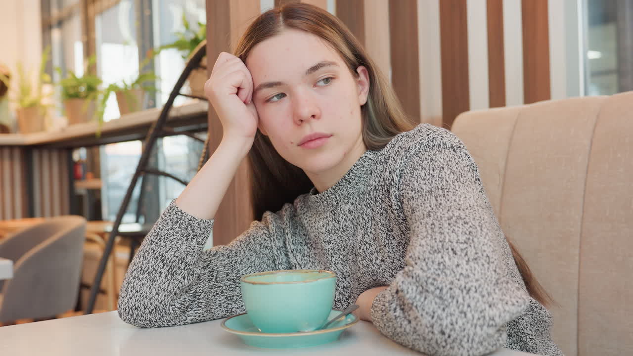 Elegant lady seated in lunch room resting arm on table while supporting head with other hand, gazing thoughtfully through window with cup and spoon in front amid warm ambient cafe setting