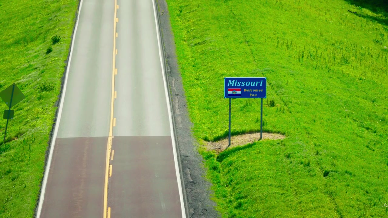 Cartello di confine di stato "Il Missouri vi dà il benvenuto" con stemma e bandiera dello stato lungo la vuota autostrada rurale US 62 vicino a Charleston, Missouri
