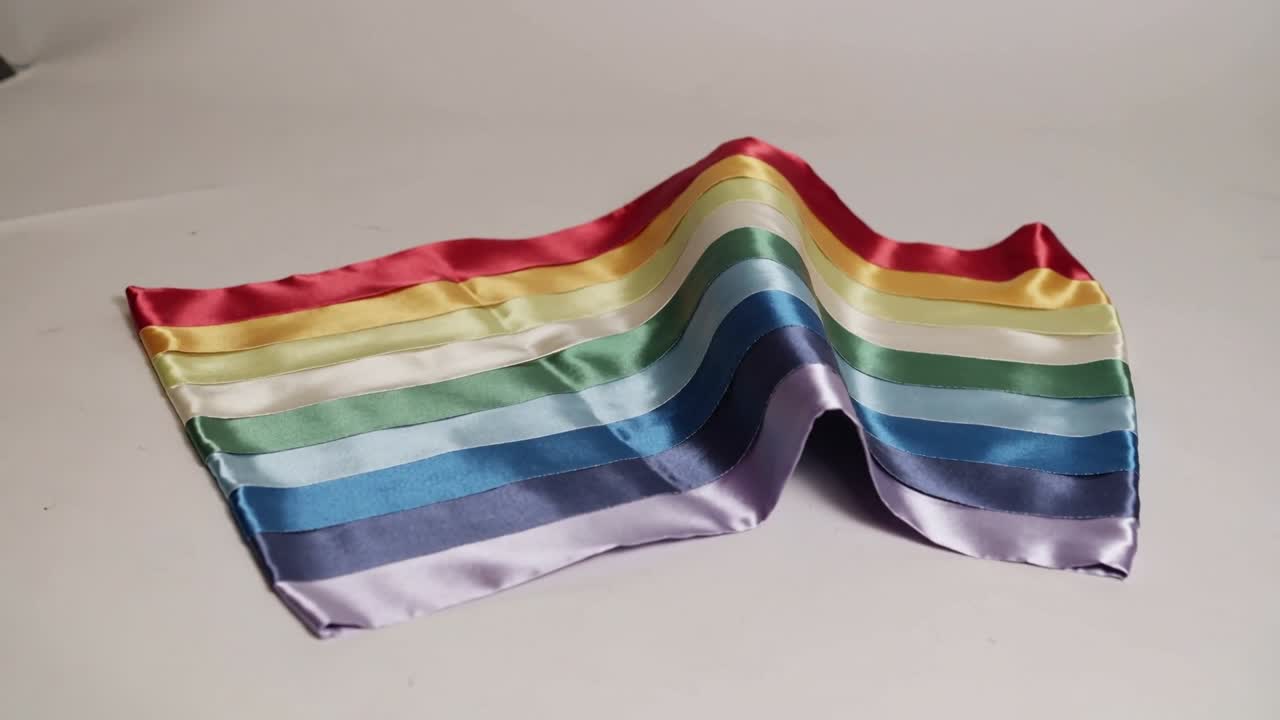 Shifting rectangular satin scarf forming peaked ridge on light tabletop, driven by soft air current