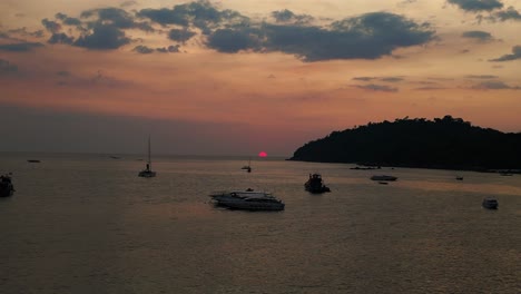 Die-Sonne-Geht-über-Dem-Ruhigen-Ozean-Unter-Und-Wirft-Einen-Warmen-Schein-Auf-Boote-Und-Eine-Insel-In-Der-Ferne,-Während-Der-Himmel-In-Orange--Und-Rosatönen-Getaucht-Wird.