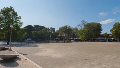 Toma-Panorámica-De-Un-Pequeño-Bote-Saliendo-Del-Puerto-Con-Autos-Pasando-Por-Ioannina