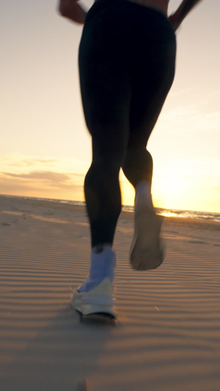 Cinematic vertical shot of an athletic woman in black tights and white shoes running along a sandy beach at golden hour, capturing coastal fitness and a healthy lifestyle