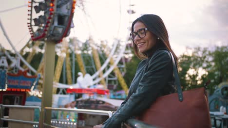 joven elegante con gafas sonriendo alegremente frente a una rueda de la fortuna en un parque de atracciones
