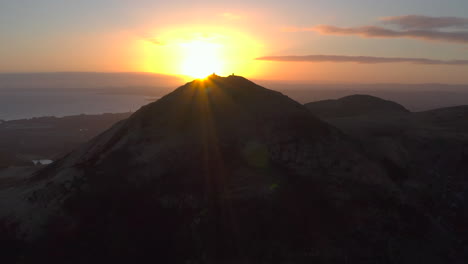 Aerial-view-of-the-sun-rising-behind-Arthur's-Seat-in-the-Holyrood-Park,-Edinburgh,-Scotland