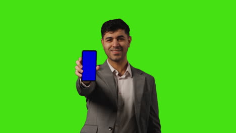 smiling businessman holding blue screen mobile phone towards camera standing against green screen background